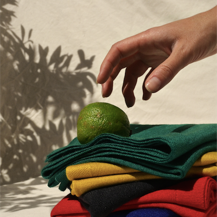 Hand reaching out to 7 folded cashmere knits in various colors. A random lime is resting on top of the cashmere. There is a shadow of foliage in the background.  . 