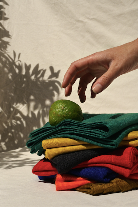 Hand reaching out to 7 folded cashmere knits in various colors. A random lime is resting on top of the cashmere. There is a shadow of foliage in the background.  . 