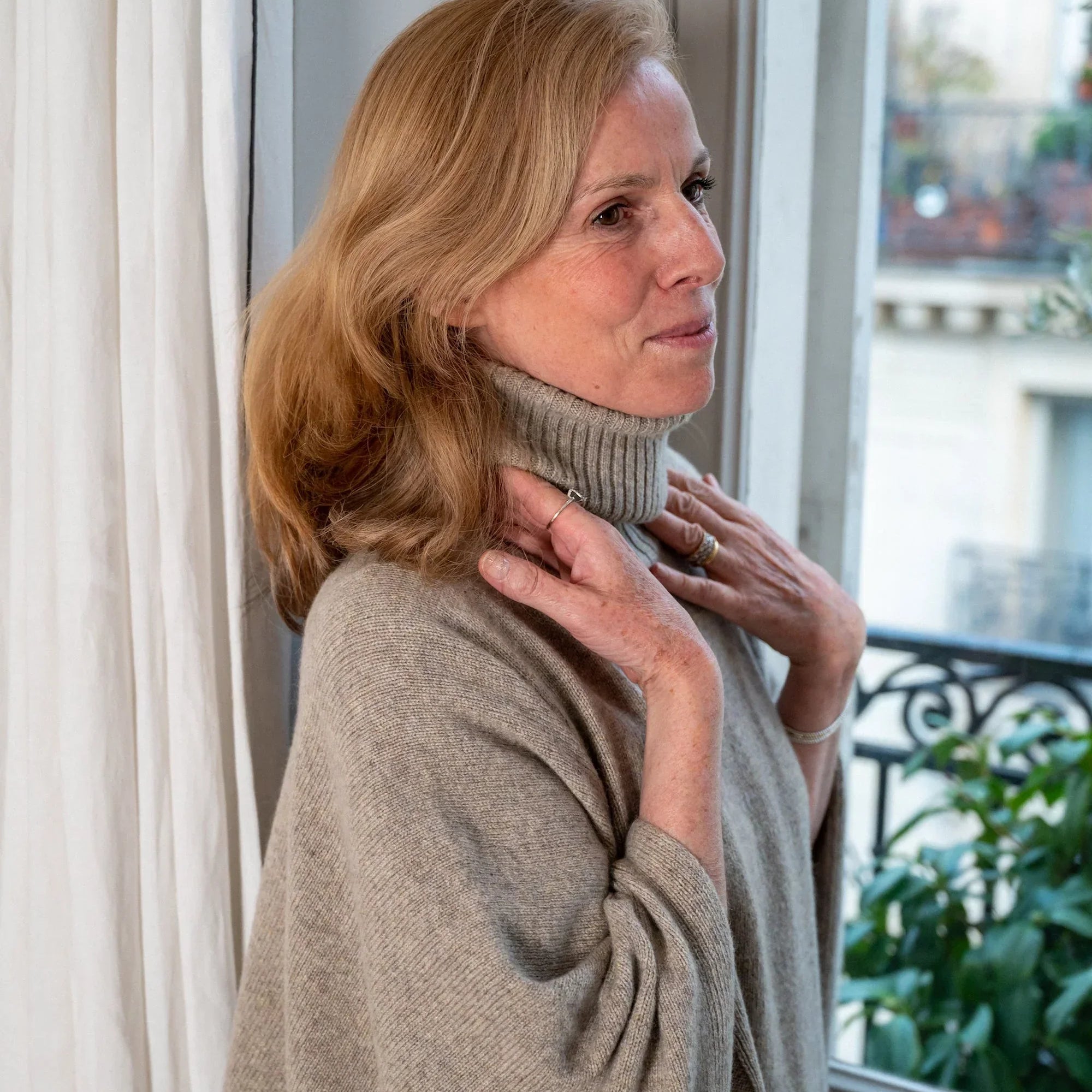 Adélaïde d’Andigné wearing a light grey turtleneck cashmere sweater by Linnea Lund, standing by the window in her Paris apartment.