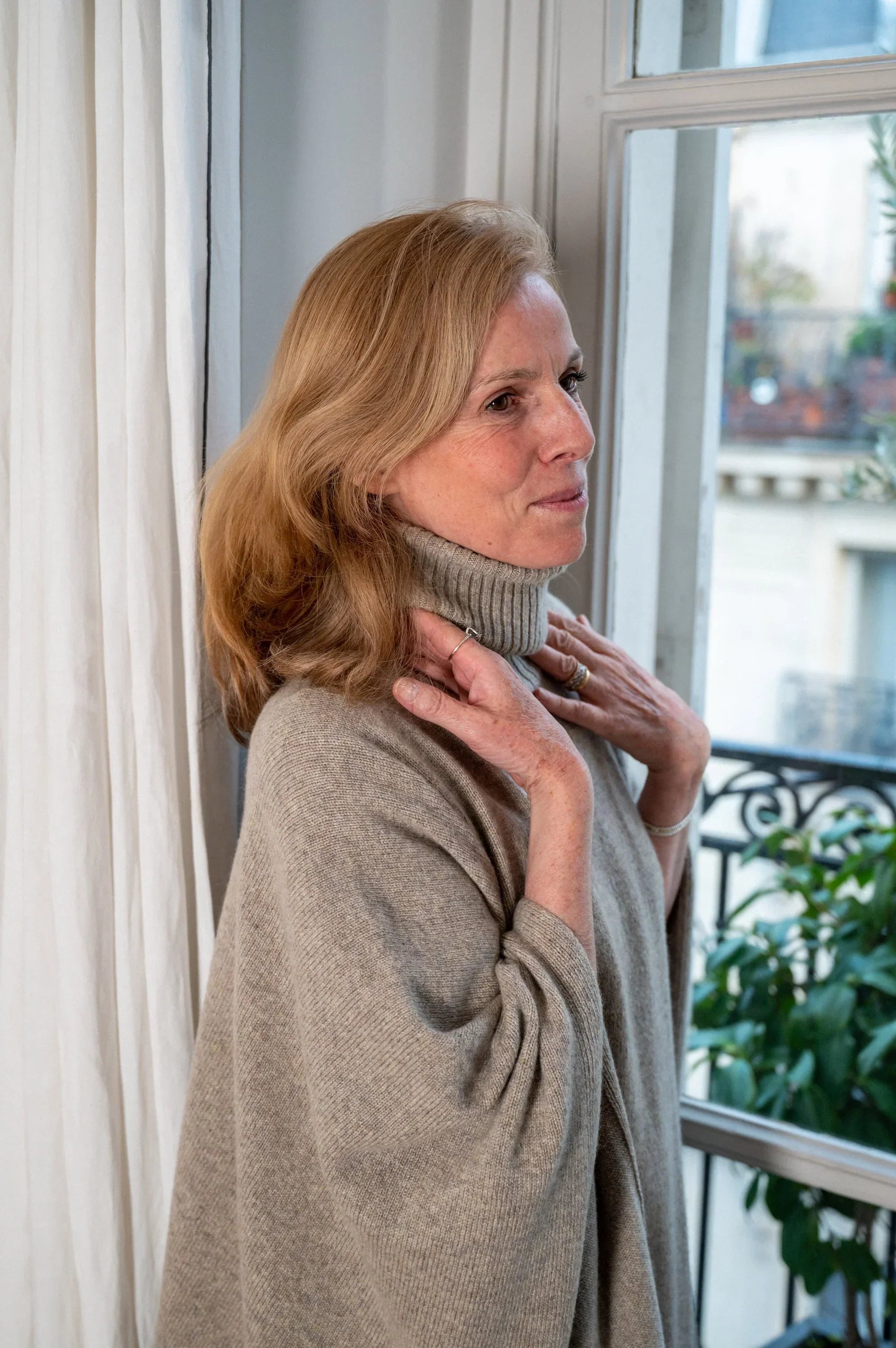 Adélaïde d’Andigné wearing a light grey turtleneck cashmere sweater by Linnea Lund, standing by the window in her Paris apartment.