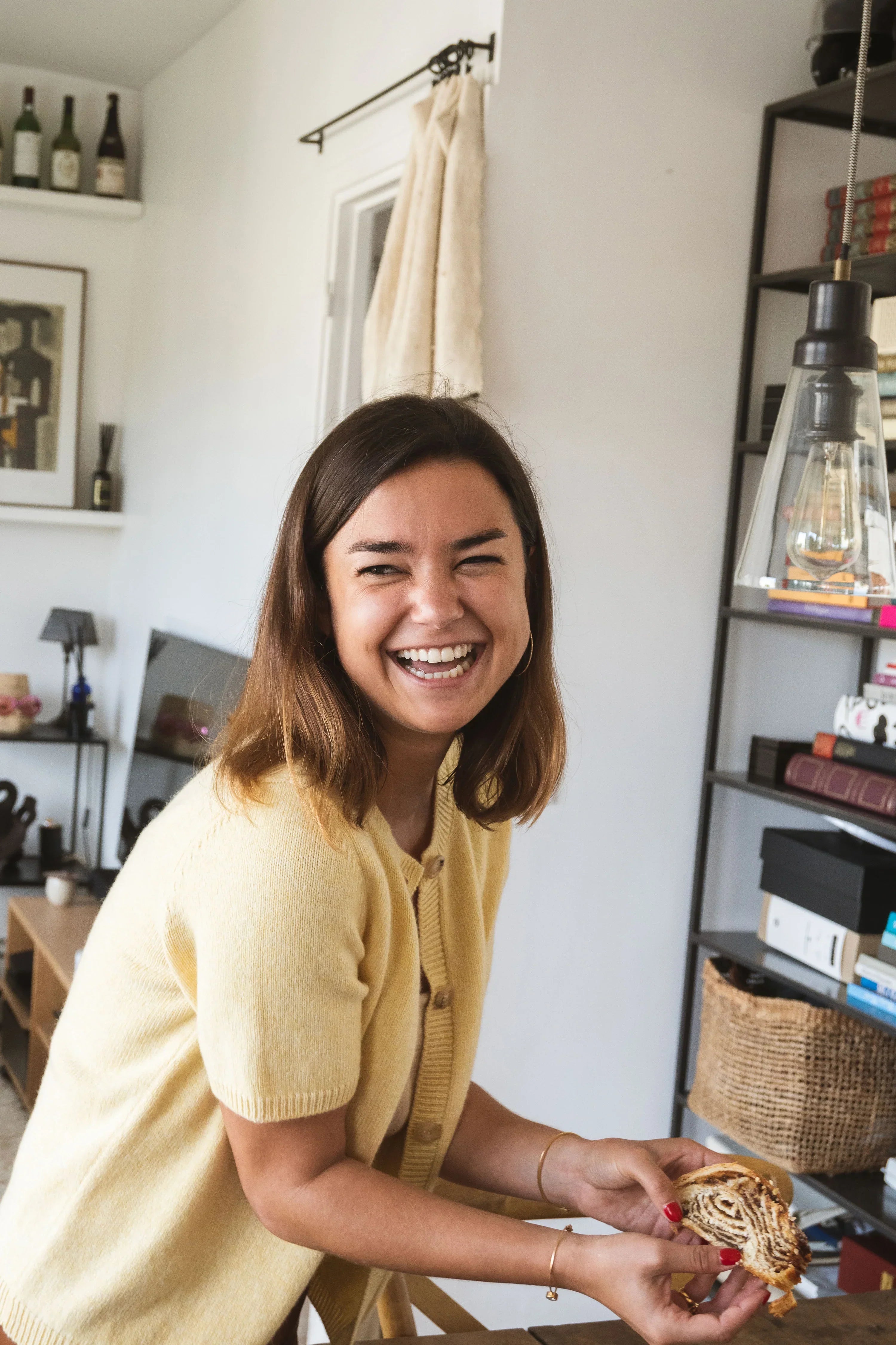 Jessica Troisfontaine wearing Linnea Lund’s Clara cardigan in honey cashmere, smiling warmly while enjoying a Swedish cinnamon roll in her Paris apartment.