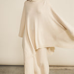 A woman look wearing an oversized white turtleneck cashmere poncho with cut-sides, paired with white pant | Angela | Linnea Lund