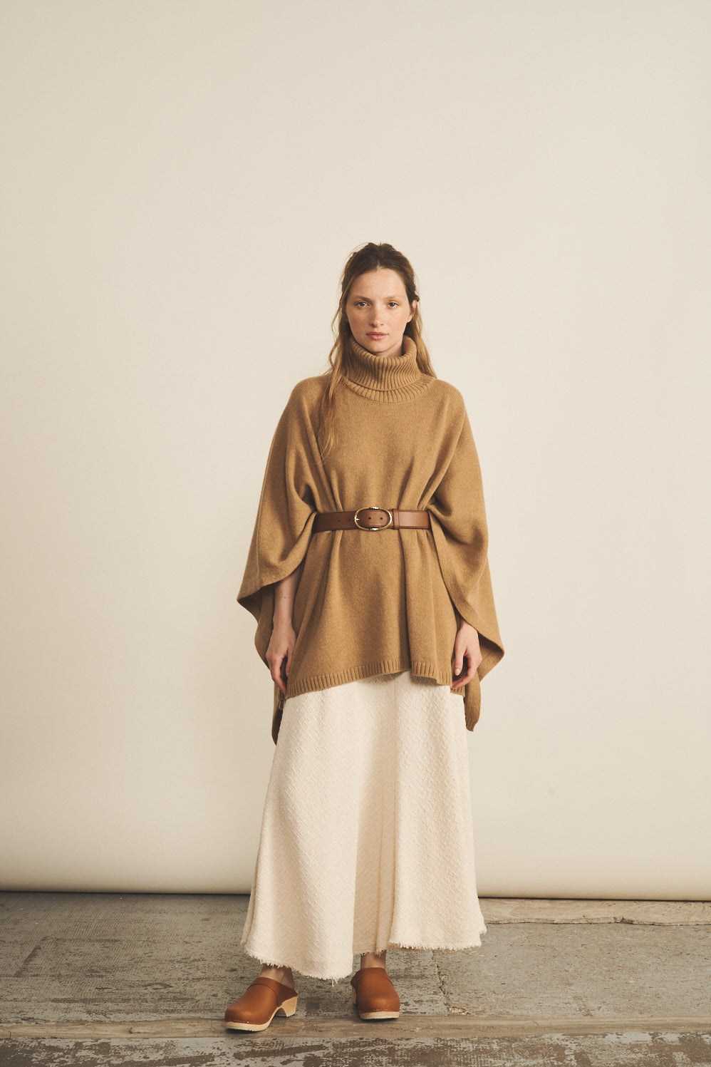 A woman look wearing an oversized camel turtleneck cashmere poncho with cut-sides, paired with white skirt | Angela | Linnea Lund